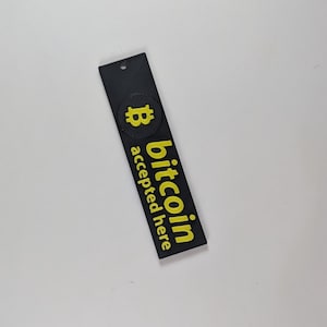 May include: A black rectangular tag with a yellow Bitcoin logo and the words "bitcoin accepted here." The tag has a small hole at the top. The Bitcoin logo is a yellow "B" inside a yellow circle.