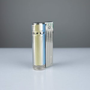 Puede incluir: Un encendedor plateado con un diseño a rayas amarillo y azul. El encendedor tiene una superficie texturizada y una pequeña ventana redonda en el lateral.