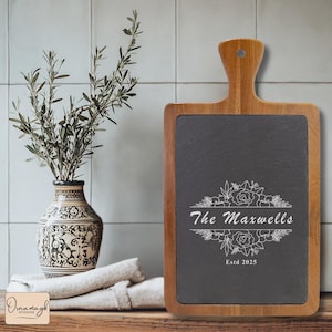 May include: Wooden serving board with a dark gray slate center. The board features the text "The Maxwells" and "Estd 2025" in white, surrounded by a floral design. The board has a handle and is displayed on a wooden surface.