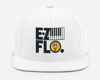 以下が含まれることがあります： 白いフラットブリムの野球帽。「EZ FLO」の文字が黒いブロック体で、ピアノの鍵盤のグラフィックと、黄色の円の中のマイクのアイコンが特徴です。