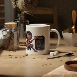 Könnte beinhalten: Weiße Keramiktasse mit einer Cartoon-Illustration einer Person, die vor einem Verstärker Bass spielt. Die Person hat braune Haut, braunes Haar und trägt Kopfhörer.