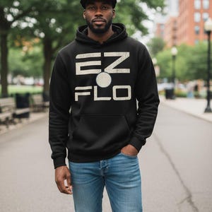 Op de afbeelding: Zwarte hoodie met de tekst "EZ FLO" in een verweerde witte letter. De persoon op de foto draagt een zwarte baseballcap met een witte "b" en een blauwe jeans. De achtergrond toont een straat met bomen.