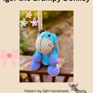 May include: A handmade blue, purple, and white crochet donkey plush toy. The text "Igor the Grumpy Donkey" is at the top, and "Pattern by S&H Handmade" is at the bottom. The donkey has large purple feet and a grumpy expression.
