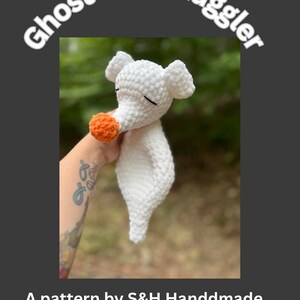 May include: A white crocheted Ghost Dog Snuggler toy with an orange nose and black stitched eyes. The text "Ghost Dog Snuggler" is arched above the toy. The text "A pattern by S&H Handdmade" is below the toy.