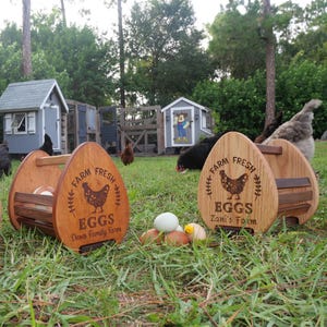 May include: Wooden egg-shaped egg holders with handles, engraved with "Farm Fresh Eggs" and a hen illustration. The holders are personalized with farm names. Several eggs are visible on the grass, with chicken coops in the background.
