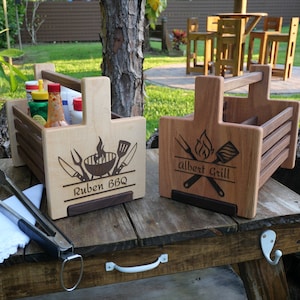 May include: Two wooden grilling caddies with handles. The caddy on the left is light wood with the text "Ruben BBQ" and a grill, spatula, and knives. The caddy on the right is dark wood with the text "Albert Grill" and a grill, spatula, and forks.