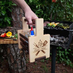 K&ouml;nnte beinhalten: Grill-Kadette aus Holz mit Griff, drei F&auml;chern f&uuml;r Flaschen und einem unteren Regal. Sie hat ein eingebranntes Design mit der Aufschrift "Dad's BBQ" mit einer Flamme, einem Spatel und einer Gabel.