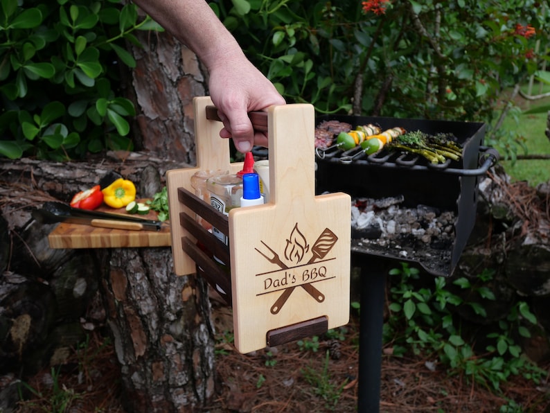 K&ouml;nnte beinhalten: Grill-Kadette aus Holz mit Griff, drei F&auml;chern f&uuml;r Flaschen und einem unteren Regal. Sie hat ein eingebranntes Design mit der Aufschrift "Dad's BBQ" mit einer Flamme, einem Spatel und einer Gabel.