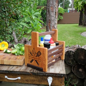 K&ouml;nnte beinhalten: Ein Grill-Caddy aus Holz mit Griff, personalisiert mit "The Espinoza Family". Er hat ein Spatel- und Gabeldesign mit Flammen. Der Caddy enth&auml;lt Flaschen mit Gew&uuml;rzen und Saucen.