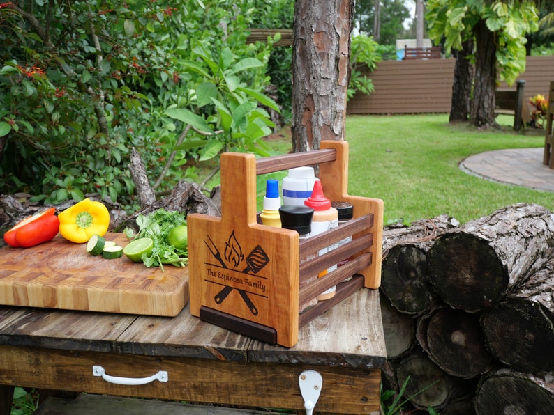 K&ouml;nnte beinhalten: Ein Grill-Caddy aus Holz mit Griff, personalisiert mit "The Espinoza Family". Er hat ein Spatel- und Gabeldesign mit Flammen. Der Caddy enth&auml;lt Flaschen mit Gew&uuml;rzen und Saucen.