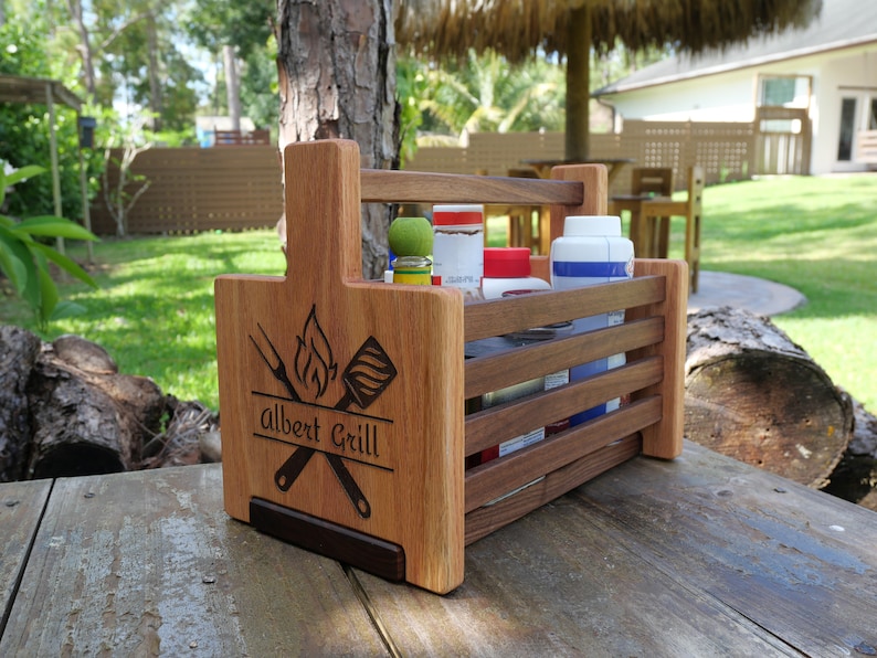 K&ouml;nnte beinhalten: Ein Grill-Caddy aus Holz mit einem Griff, personalisiert mit dem Namen "Albert Grill" und einem Design aus einem Spatel und einer Gabel, die &uuml;ber einer Flamme gekreuzt sind. Der Caddy hat vier F&auml;cher zum Verstauen von Grillwerkzeug und Gew&uuml;rzen.