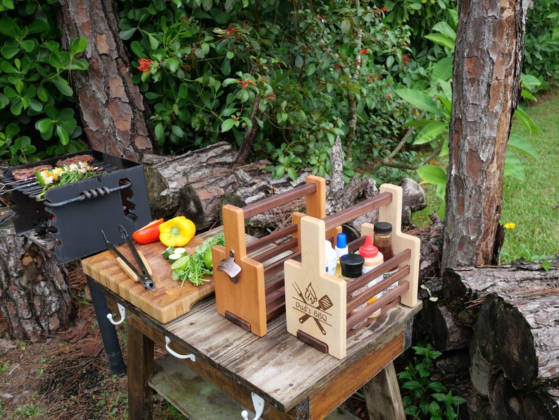 K&ouml;nnte beinhalten: Zwei Grill-Caddies aus Holz mit Griffen. Der Caddy rechts hat den Text "Dad's BBQ" auf der Vorderseite eingraviert. Beide Caddies haben Flaschenhalter und einen integrierten Flaschen&ouml;ffner.