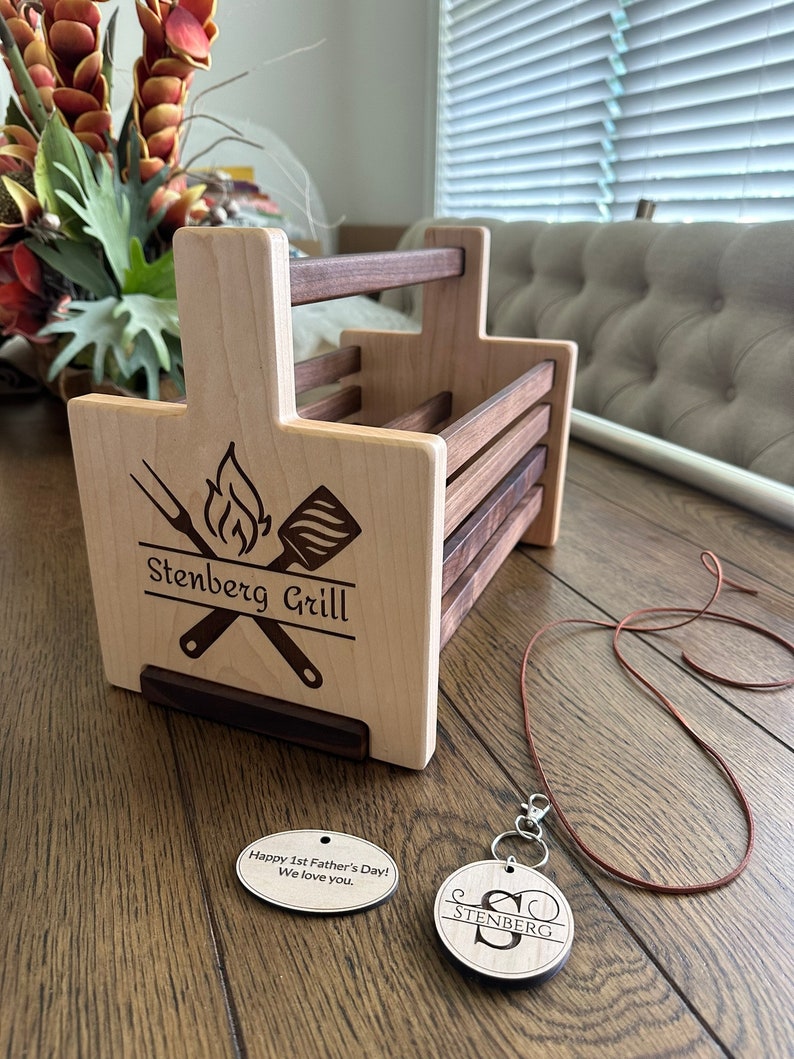 K&ouml;nnte beinhalten: Personalisierter Grillwerkzeug-Halter aus Holz mit dem Text "Stenberg Grill", der auf die Vorderseite graviert ist. Er wird mit einem passenden Holzschl&uuml;sselanh&auml;nger mit dem Text "Stenberg" und einer Lederschnur geliefert.