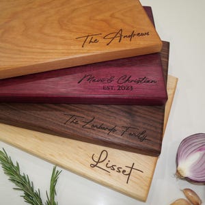 May include: A stack of rectangular wooden cutting boards in various wood tones, including light, medium, and dark shades. Each board is engraved with a different name or family name in a cursive font. Fresh rosemary, garlic cloves, and a sliced red onion are in the foreground.