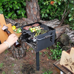 K&ouml;nnte beinhalten: Eine Grillstation aus Holz mit Flaschen&ouml;ffner, Gew&uuml;rzbeh&auml;ltern und einem Schneidebrett. Die Station steht neben einem schwarzen Holzkohlegrill mit Spie&szlig;chen mit Fleisch und Gem&uuml;se, die darauf garen. Der Grill steht auf einem Metallgestell und ist von gr&uuml;nen Pflanzen und Baumst&auml;mmen umgeben.