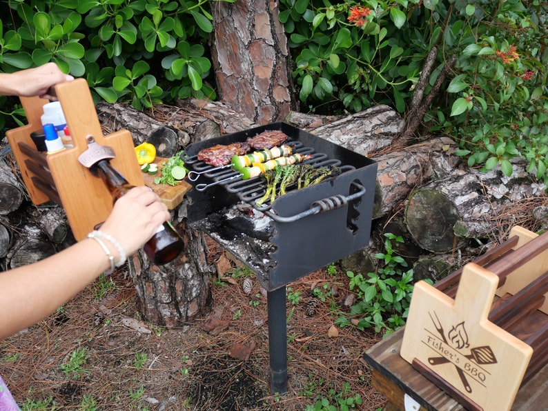 K&ouml;nnte beinhalten: Eine Grillstation aus Holz mit Flaschen&ouml;ffner, Gew&uuml;rzbeh&auml;ltern und einem Schneidebrett. Die Station steht neben einem schwarzen Holzkohlegrill mit Spie&szlig;chen mit Fleisch und Gem&uuml;se, die darauf garen. Der Grill steht auf einem Metallgestell und ist von gr&uuml;nen Pflanzen und Baumst&auml;mmen umgeben.
