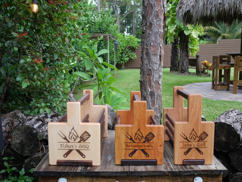 K&ouml;nnte beinhalten: Drei Grillwerkzeug-Halter aus Holz mit personalisierten Gravuren. Der erste Halter sagt "Fisher's BBQ", der zweite sagt "The Espinoza Family" und der dritte sagt "John's Grill".