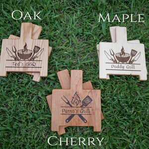 K&ouml;nnte beinhalten: Drei S&auml;tze Grilluntersetzer aus Holz, jeweils mit einer anderen Holzart: Eiche, Ahorn und Kirsche. Jeder Untersetzersatz verf&uuml;gt &uuml;ber einen personalisierten Namen und ein Grill-Design mit Grill, Spatel, Zange und Messer.
