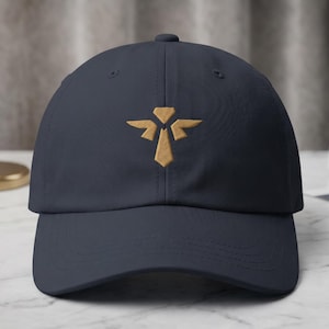 Könnte beinhalten: Eine marineblaue Baseballkappe mit einem mittig platzierten, goldfarbenen Stickemblem. Das Design besteht aus einer vertikalen Rautenform über einem nach unten zeigenden Dreieck, flankiert von zwei symmetrischen, flügelartigen geometrischen Formen.