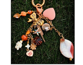 May include: A gold-coloured keyring adorned with diverse charms: a pink heart, a bow, a seashell, and a "Sweet Heart" charm. The keyring also features orange and clear beads, a clear crystal ball, and a white and red stone pendant.