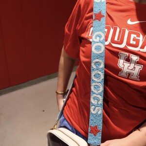 May include: A light blue beaded purse strap with the words "GO COOGS" in white. The strap has red star accents. The person in the image is wearing a red t-shirt with the word "COUGARS" and a logo.