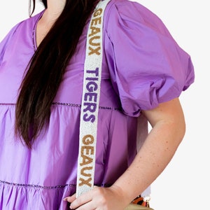 May include: A white beaded bag strap with the words "TIGERS GEAUX" in purple and gold lettering.