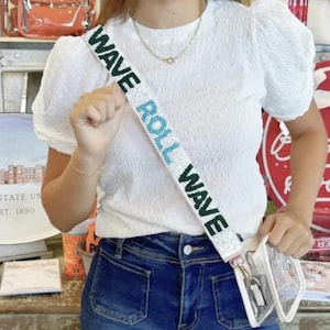 May include: A white textured short-sleeve top with a beaded crossbody bag strap featuring the text WAVE ROLL WAVE in dark green and light blue beads on a white background, attached to a clear plastic bag with gold-tone metal hardware.
