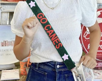 Może przedstawiać: Zielony, koralikowy pasek na ramię z napisem "GO CANES" z czerwonych koralików. Pasek ma różowe gwiazdki i jest połączony z przezroczystą torbą. Osoba ma na sobie białą górę i niebieskie dżinsy.