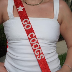 May include: A red sash with the words "GO COOGS" in white, beaded letters. A white star is at the top of the sash. The sash is worn over a white tank top. A gold chain necklace is also visible.