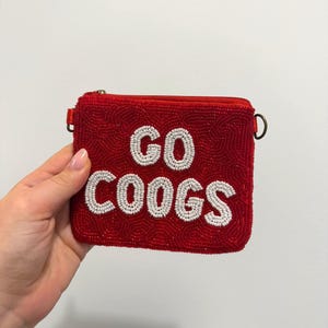 May include: A rectangular red beaded pouch featuring the white beaded text GO COOGS on the front. The pouch has a top zipper closure and a small metal D-ring on the side for attachment. The entire surface is covered in small, uniform red beads.