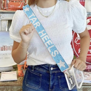 May include: A light blue beaded bag strap with white lettering that reads "TAR HEELS" and a white star. The strap is worn over a white t-shirt and blue jeans.