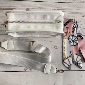 May include: A white clear crossbody bag with a gold zipper and a white adjustable strap. The bag is paired with a pink and white patterned scarf with nautical accents.