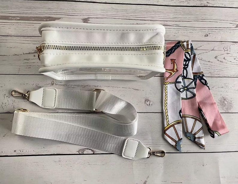 May include: A white clear crossbody bag with a gold zipper and a white adjustable strap. The bag is paired with a pink and white patterned scarf with nautical accents.