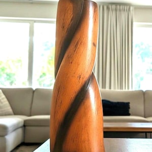 May include: A tall, cylindrical wooden vase featuring a distinct spiral or twisted design carved into the body. The wood displays a warm, light brown natural grain with darker, contrasting brown streaks following the spiral grooves of the structure.