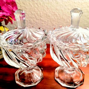 May include: Two clear glass candy dishes with lids, featuring a swirling ribbed pattern on the bowls and matching lids. Each dish stands on a hexagonal pedestal base, crafted from transparent glass with a reflective, polished finish throughout.