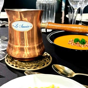 May include: A copper sauce pot with a long wooden handle and a white oval label featuring the text Le Saucier in blue script. The pot sits on a round, ornate gold-toned metal trivet, showcasing a polished metallic finish and a sturdy design.