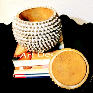May include: A decorative, spiky, white and gold-colored ceramic bowl with a matching lid. The bowl is sitting on top of a stack of books with the words "Art Deco" visible on the cover. The background is black.
