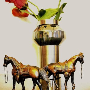 May include: Two bronze-colored horse figurines with detailed saddles and bridles featuring hanging chains, positioned in front of a cylindrical metallic vase containing a bouquet of red and orange tulips with green stems and leaves.