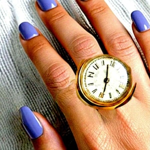 May include: A gold-toned ring featuring a round, clock-face design with a white dial, black Roman numerals, and black hands. The watch face is set within a polished gold metal frame, creating a prominent, circular statement piece on the finger.