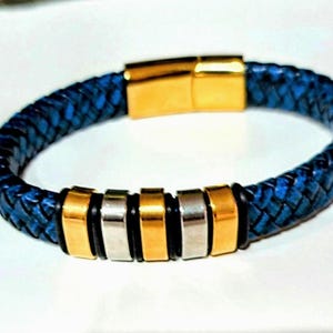 May include: A braided blue leather bracelet featuring a gold-toned metal clasp and a series of alternating gold and silver-toned metal rectangular beads separated by thin black rings along the front section of the band.