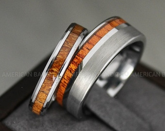 May include: Two wedding bands, one with a silver finish and the other with a wood inlay. The wood inlay is a light brown color with darker brown stripes. The bands are on a gray surface.