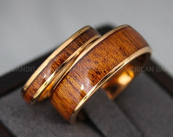 May include: Two gold-toned wedding bands with a brown wood inlay. The bands are stacked on top of each other.