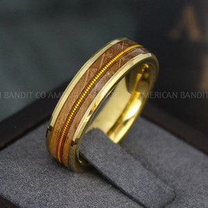 May include: A gold tungsten carbide ring with a brown wood inlay and a thin gold band. The ring is in a black box.