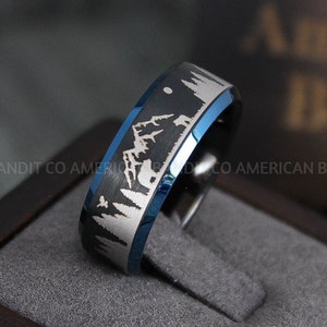May include: A blue and silver tungsten carbide ring with a mountain scene engraved on the band. The ring is in a wooden box.