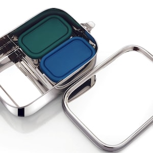 May include: Stainless steel lunch box with two compartments, each with a colored silicone lid. One lid is green, the other is blue. The lid is separate from the box. The lunch box has a shiny, metallic finish.