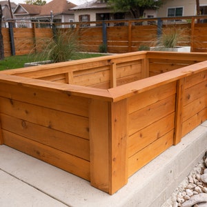 Puede incluir: Una jardinera de madera grande, rectangular, con un acabado de madera natural. La jardinera está hecha de tablones de cedro y tiene un borde elevado alrededor de la parte superior.