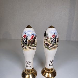 May include: Two white ceramic beer tap handles with a gold base. Each handle features a hunting scene with riders in red coats and the text "Ye Olde Pub handle".