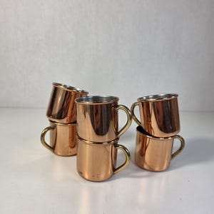 Może przedstawiać: Zestaw pięciu małych miedzianych kubków z mosiężnymi uchwytami, ułożonych w stos. Każdy kubek ma polerowane metaliczne miedziane wykończenie zewnętrzne oraz gładką, srebrzystą wyściółkę wewnętrzną, z zakrzywionym uchwytem przymocowanym z boku.