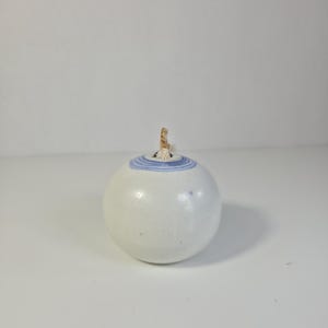 May include: A white ceramic oil lamp with a spherical shape. The lamp features a blue and white striped design around the top and a natural fiber wick. The lamp is designed for aromatherapy or decorative purposes.