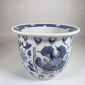 Peut inclure: Un cache-pot en céramique blanche doté d'un bord évasé et d'un corps arrondi. L'extérieur est orné de motifs floraux et de feuilles bleus, incluant de grandes fleurs de lotus et du feuillage, avec un motif de pétales répété sur le bord.